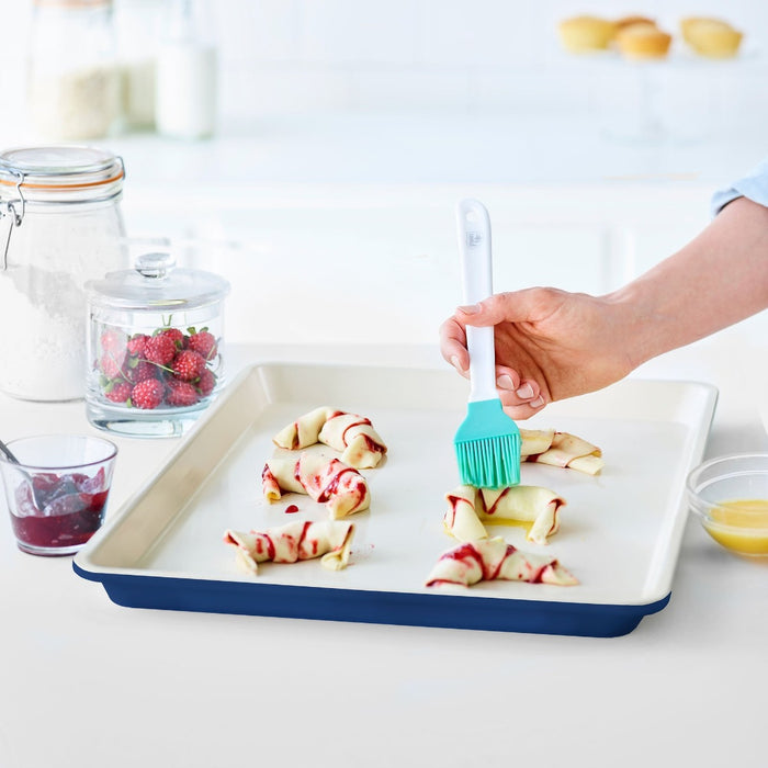 Hand brushing pastry on a navy Blue GreenLife ceramic nonstick cookie sheet with jam and strawberries next to it.