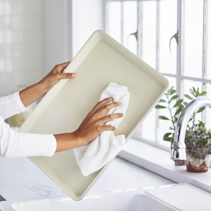 Person cleaning navy ceramic nonstick cookie sheet by a kitchen sink with green plants in the background.