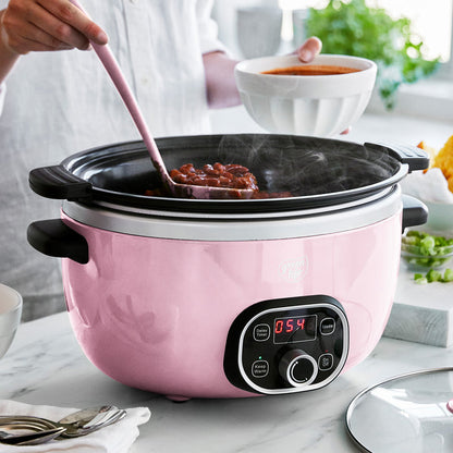 Pink slow cooker with digital display on a kitchen counter, person using a pink ladle to serve food.