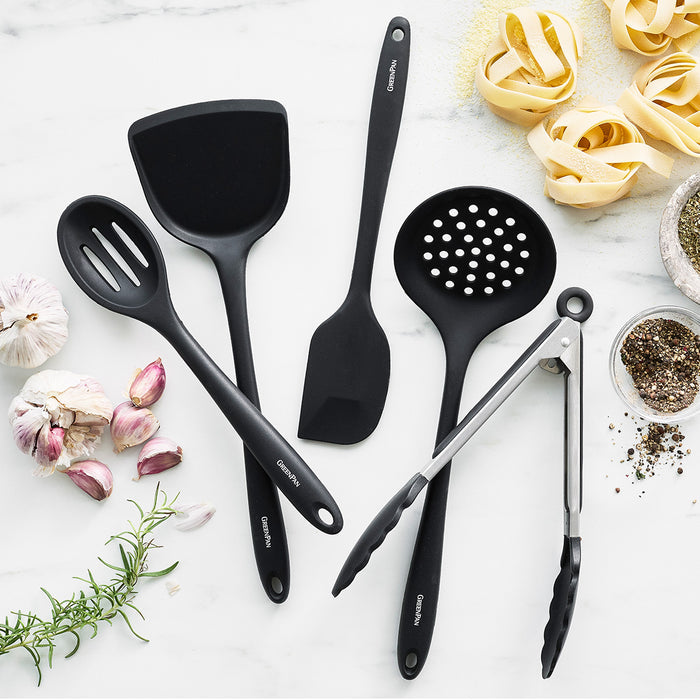 Black utensil set and white ceramic skillet with herbs, garlic, pepper, and pasta on a marble surface.
