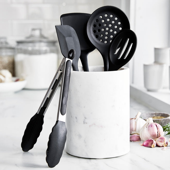 Black kitchen utensils in a marble holder on a counter next to garlic bulbs and spice jars.