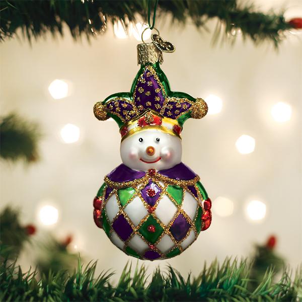 A festive Christmas tree ornament shaped like a snowman wearing a jester hat, decorated in purple, green, gold, and white, hanging among green tree branches with blurred lights in the background.