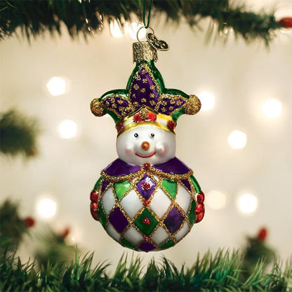 A festive Christmas tree ornament shaped like a snowman wearing a jester hat, decorated in purple, green, gold, and white, hanging among green tree branches with blurred lights in the background.