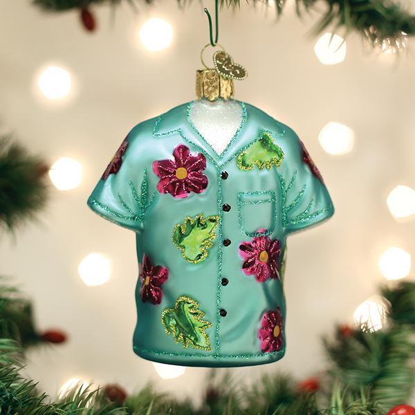A Christmas tree ornament shaped like a teal Hawaiian shirt with pink and green floral decorations hangs among green branches and blurred white holiday lights in the background.