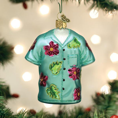 A Christmas tree ornament shaped like a teal Hawaiian shirt with pink and green floral decorations hangs among green branches and blurred white holiday lights in the background.