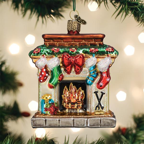 A festive Christmas tree ornament shaped like a fireplace, decorated with garland, a red bow, four colorful stockings, a fire in the hearth, and holiday gifts, hanging from a tree branch with lights in the background.
