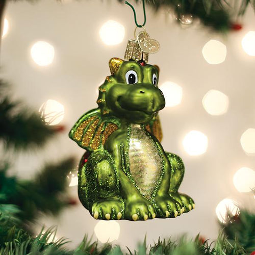 A green dragon Christmas ornament with golden wings and a sparkly belly hangs from a tree branch, surrounded by blurred festive lights in the background.