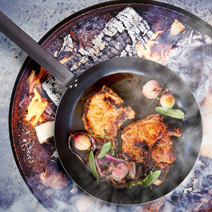 Pork chops, onions, and herbs cooking in a Merten  Storck carbon steel frypan set on a stovetop.