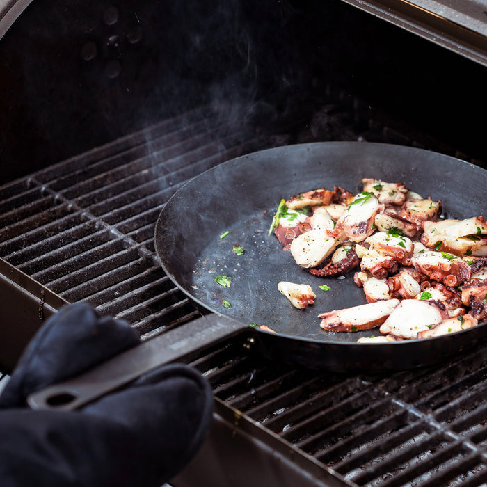 Sliced octopus and herbs being cooked in a Merten  Storck carbon steel 12-inch frypan, hands with black oven mitts.