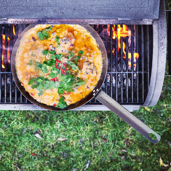 Carbon steel 12-inch frypan with soup, herbs, and chili cooking on a grill over grass.