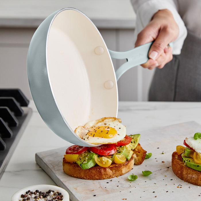 Person using a frying pan to place an egg on avocado toast on a marble countertop.