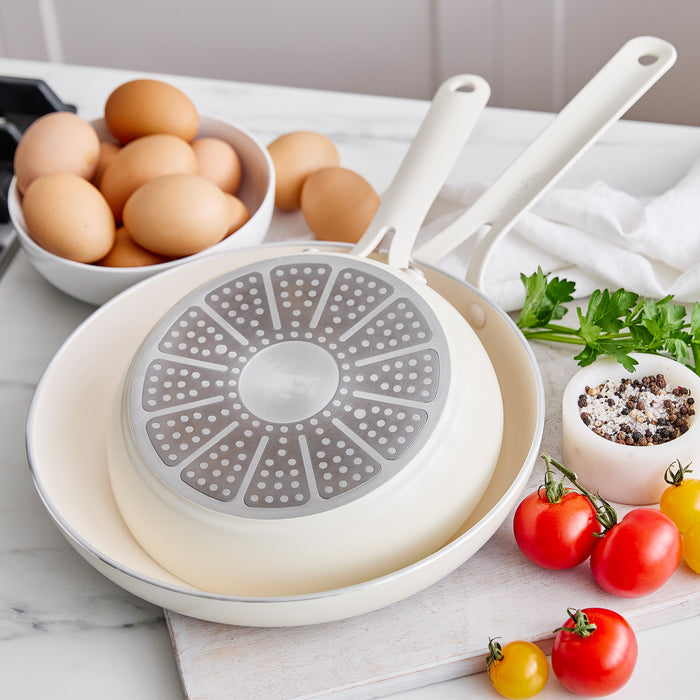 White frying pans on a kitchen counter with eggs and vegetables.