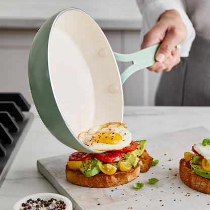 Person using a green frying pan to place an egg onto avocado toast on a marble countertop.