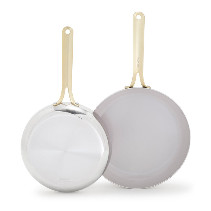 Set of two stainless steel frypans with gold handles, one face up and one face down on a white surface.