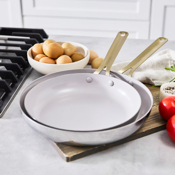 Two stainless steel frypans, 10-inch and 12-inch, on a kitchen counter beside eggs and vegetables near a stove.