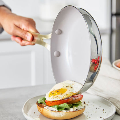 A hand slides a fried egg from a stainless steel frypan onto a bagel topped with spinach, tomato, and cream cheese.
