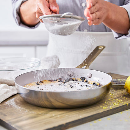 Blueberry dessert in a stainless steel frypan on a wooden board, topped with powdered sugar being sprinkled.