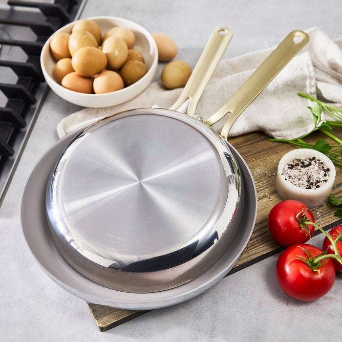 Two stainless steel frypans, 10 and 12, stacked on a board with eggs, tomatoes, herbs, and salt and pepper nearby.