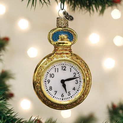 A gold pocket watch ornament showing the time as 5:00 hangs on a Christmas tree with blurred white lights in the background.