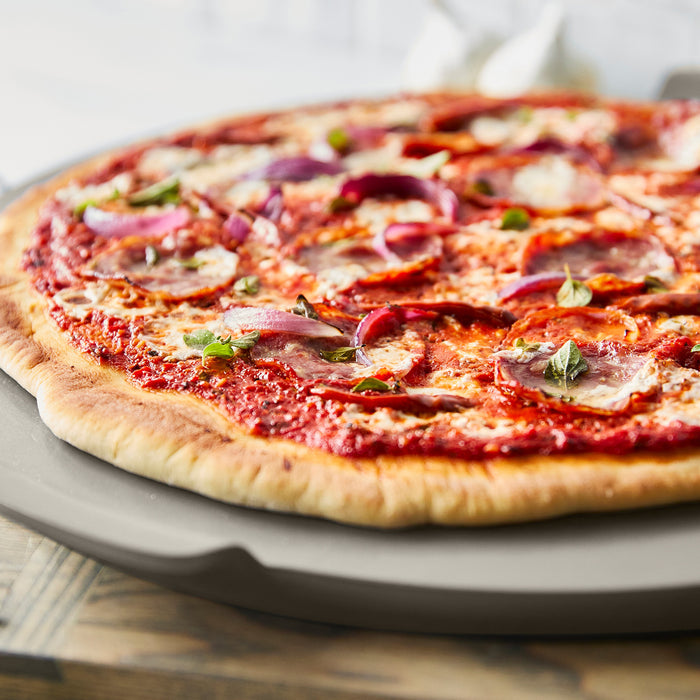 Pizza with tomato, cheese, onions, and herbs on a taupe Premiere Ovenware 16.5-inch round pizza baker.