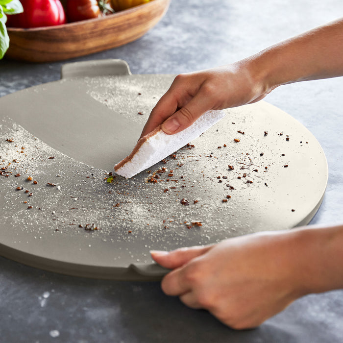 Hands brushing crumbs off a taupe 16.5-inch round pizza baker.