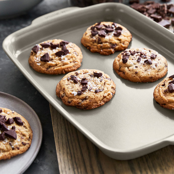 Six chocolate chip cookies with sea salt on a gray ceramic nonstick baking sheet, one cookie served on a white plate.