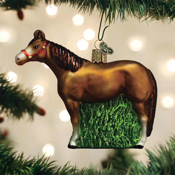 A glass Christmas tree ornament shaped like a brown horse with a red bridle, hanging among green pine branches with blurred white lights in the background.