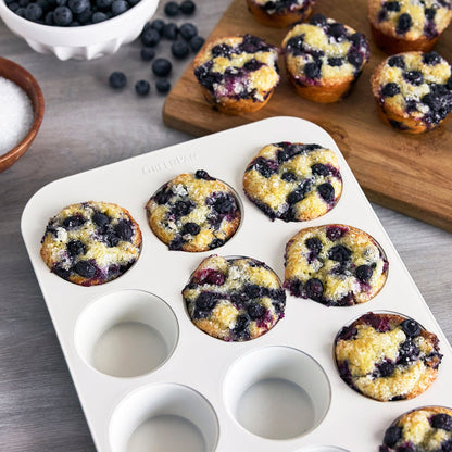 Cream 12-cup muffin pan with baked blueberry muffins next to a wooden board and small bowls of berries.