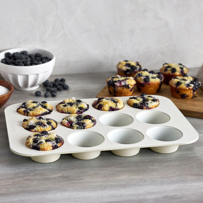 Cream-colored 12-cup muffin pan with baked blueberry muffins, extra muffins and blueberries in the background.