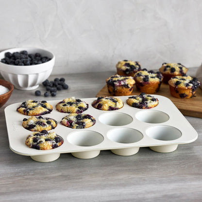 Cream-colored 12-cup muffin pan with baked blueberry muffins, extra muffins and blueberries in the background.
