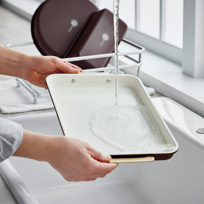 Hands rinsing a dark red Reserve Bakeware 13″ x 9″ quarter sheet pan under a kitchen faucet.