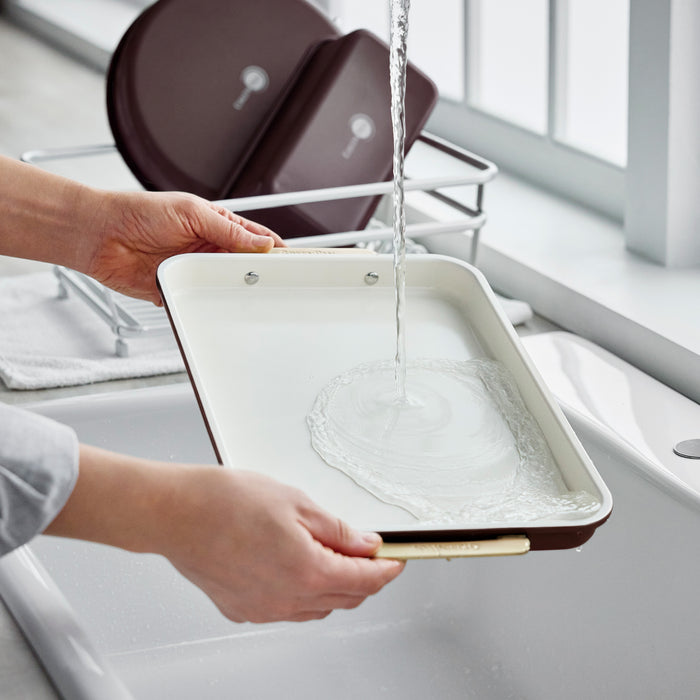 Hands rinsing a dark red Reserve Bakeware 13″ x 9″ quarter sheet pan under a kitchen faucet.