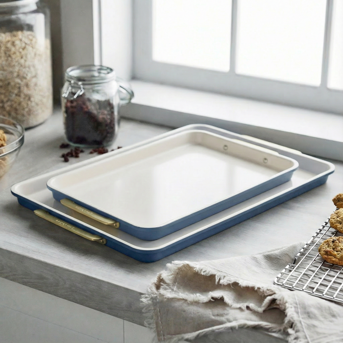 Two blue sheet pans stacked on a kitchen counter near cookies and glass jars with sunlight coming through the window.