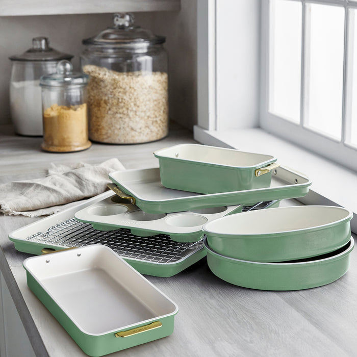 Sage green 8-piece bakeware set stacked on a kitchen counter next to glass jars and a window.
