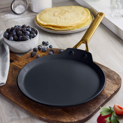 Black ceramic nonstick pan with gold handle on a wooden surface; blurred berries and crepes in the background.
