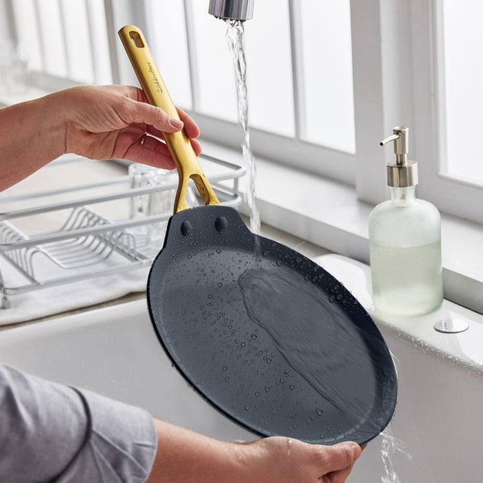 Black ceramic nonstick pan with a yellow handle being rinsed under water at a kitchen sink near a window.
