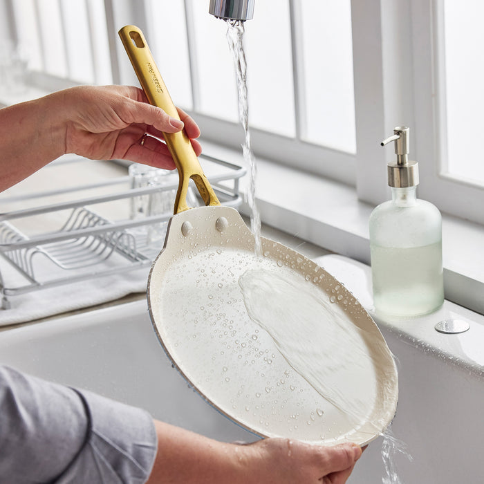 Person washing a crepe pan under running water with soap in a kitchen sink.