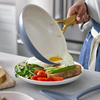 Person holding a frying pan with a cooked egg over a plate of toast, avocado, and salad.