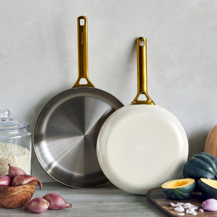 Two uncoated stainless steel frypans in cream, 10″ and 12″ sizes, on a counter with rice, onions, and vegetables nearby.