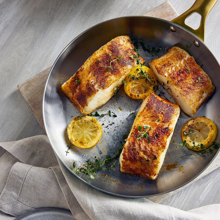 Pan-fried fish fillets with lemon slices on a pan, placed on a wooden surface.