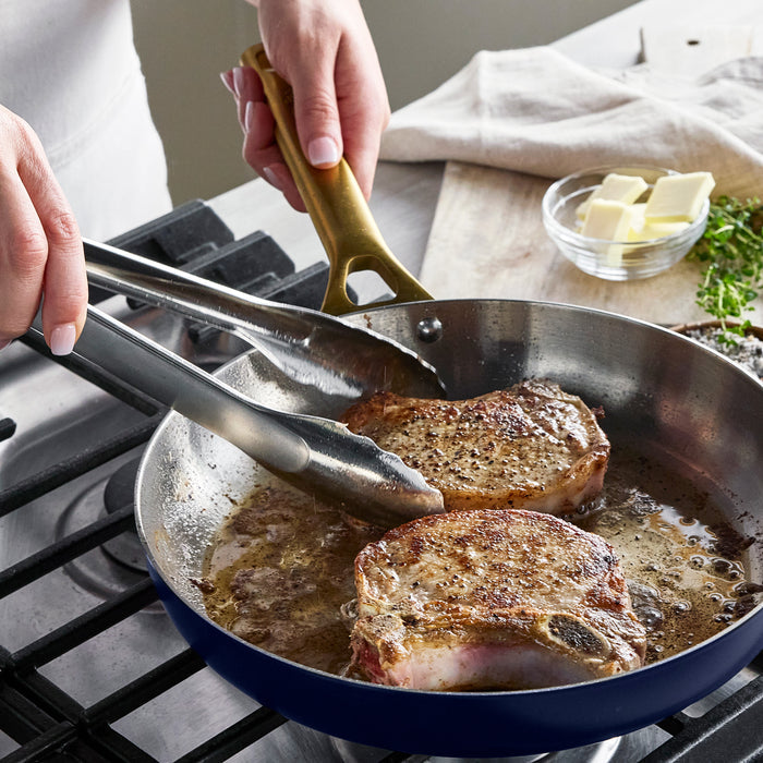 Person cooking pork chops in a pan with tongs on a stovetop.