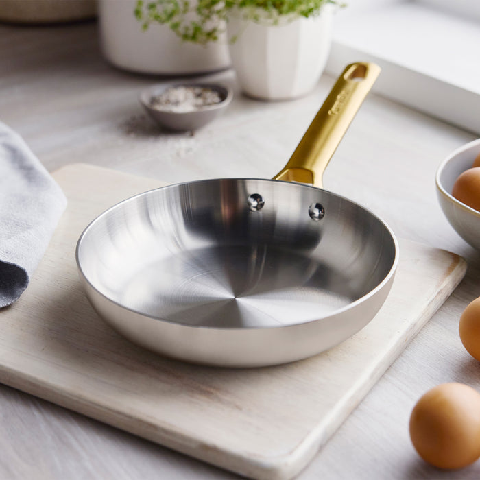 Cream uncoated stainless steel 8 frypan with a gold handle on a board, surrounded by eggs and utensils.