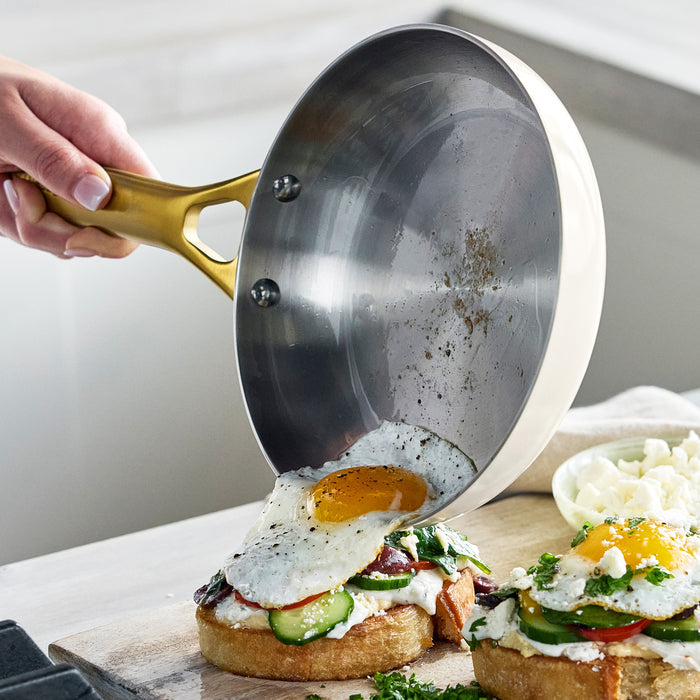 Hand pouring a sunny side up egg from an 8 cream stainless steel frypan onto veggie toast with cheese.