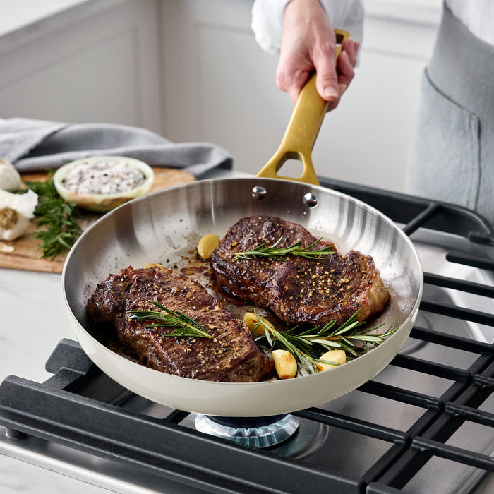 Two garlic-rosemary steaks cooking in an uncoated stainless steel frypan on a stovetop.