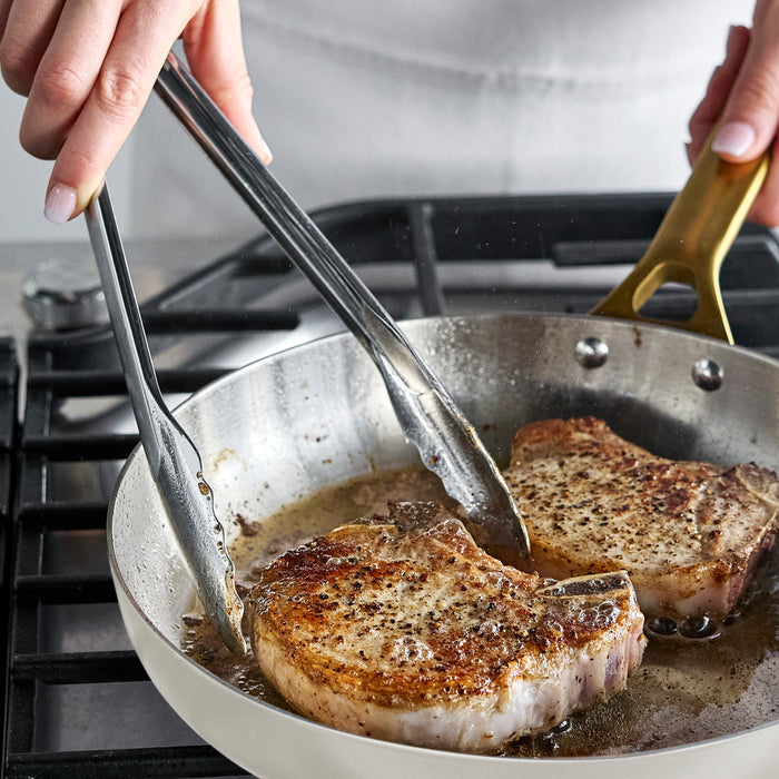 Pork chops being flipped with tongs in a cream-colored uncoated stainless steel 8 frypan on a stovetop.