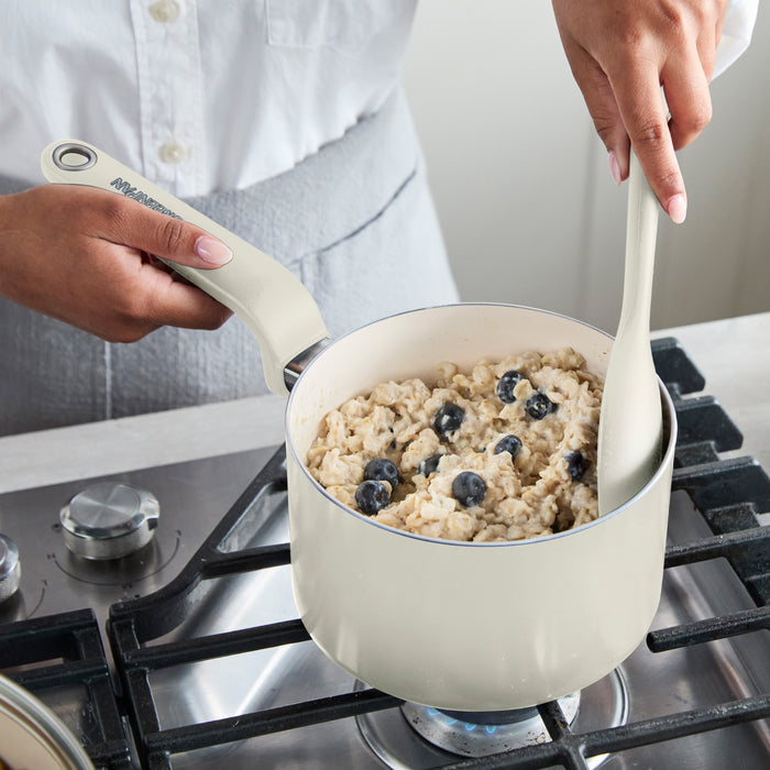 Cream ceramic saucepan with lid on a stove, blueberry oatmeal being stirred inside.