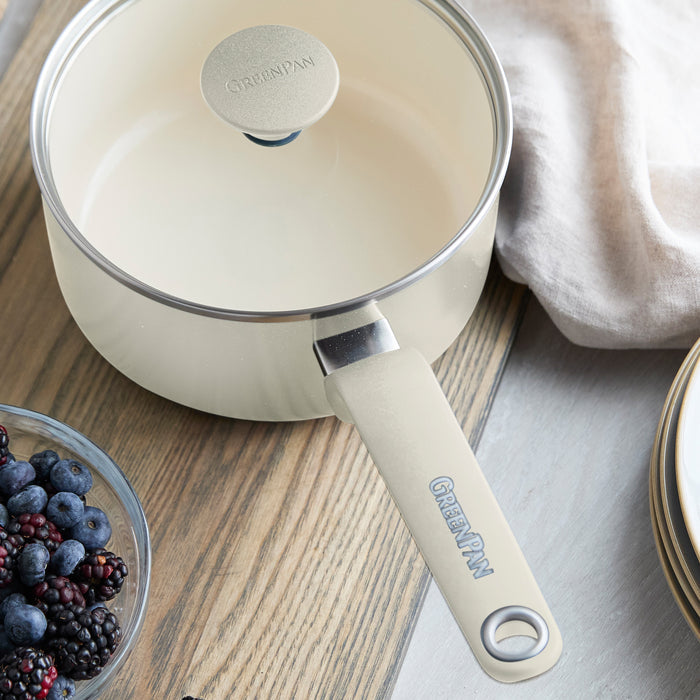 Cream ceramic 2-quart saucepan with lid on wooden table, shown beside berries, plates, and a beige napkin.
