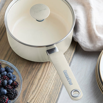 Cream ceramic 2-quart saucepan with lid on wooden table, shown beside berries, plates, and a beige napkin.