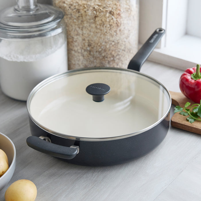 Black ceramic 5-quart sauté pan with lid on a counter next to potatoes, oats, flour, herbs, and a pepper.