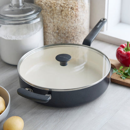 Black ceramic 5-quart sauté pan with lid on a counter next to potatoes, oats, flour, herbs, and a pepper.